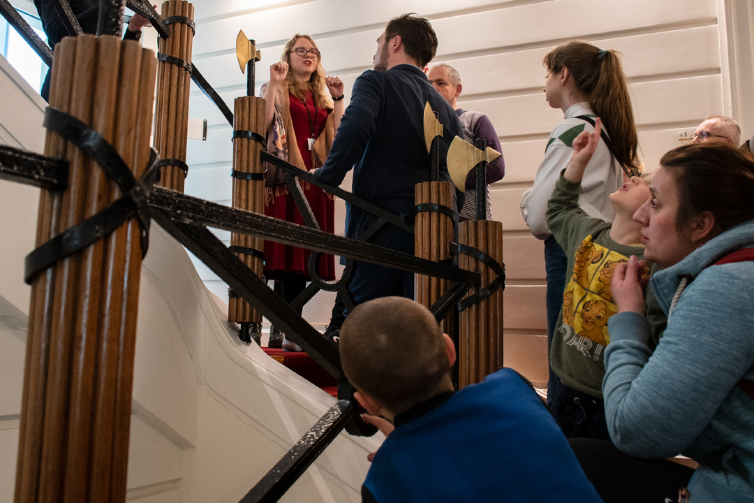 ludzie stoją na schodach w zabytkowym gmachu, słuchają ubranej w czerwona sukienkę przewodniczki