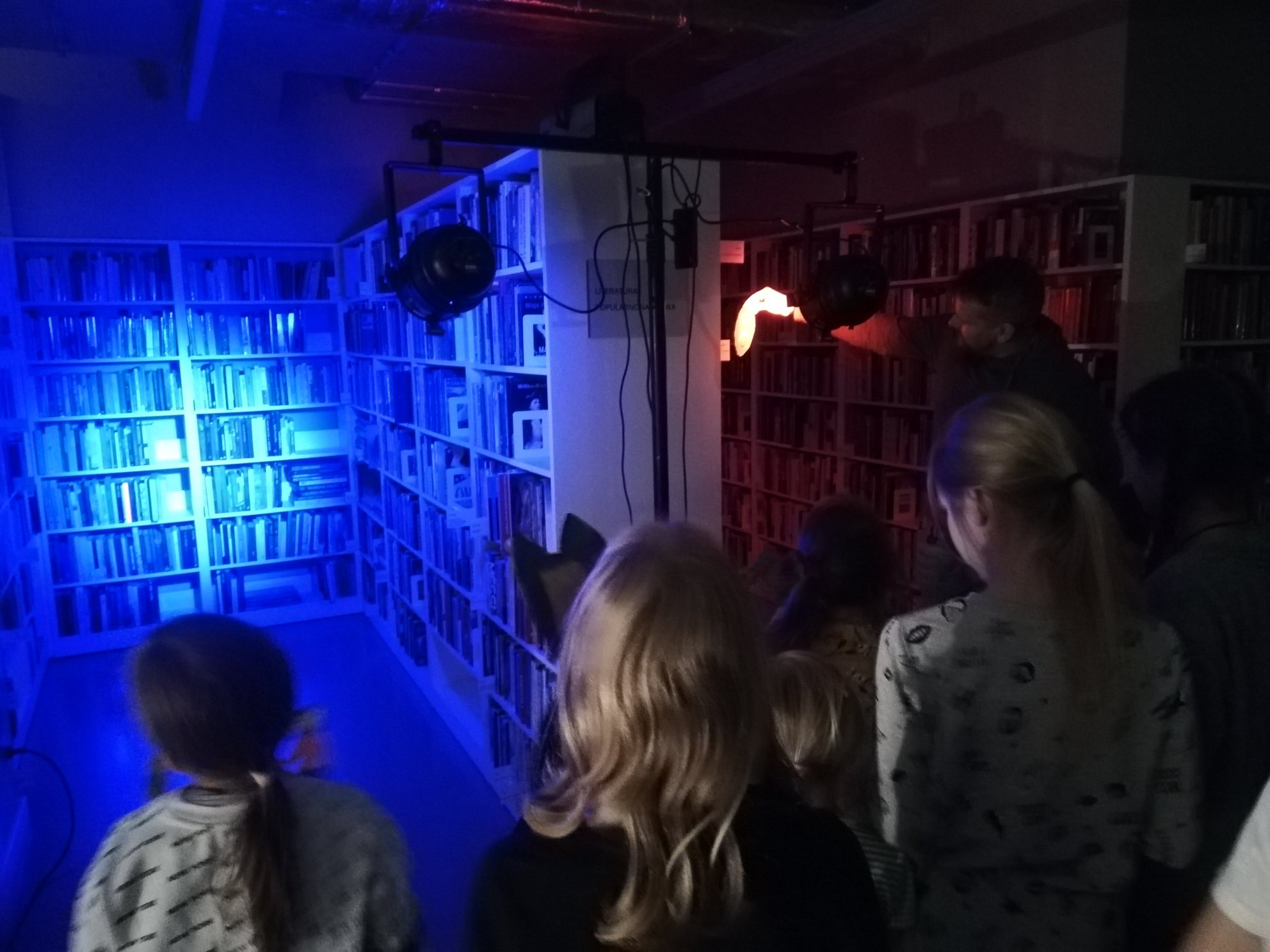 w ciemnej bibliotece widac dzieci oświetlajace regały niebieskim światłem latarki