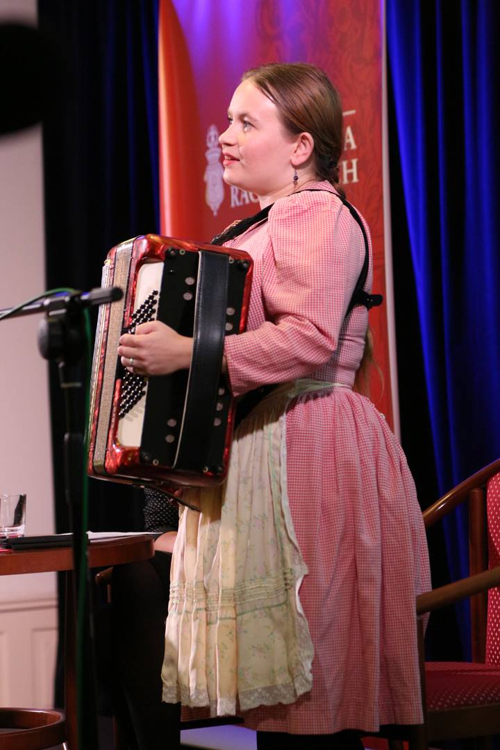 dziewczyna z akordeonem, stoi profilem, ma długi warkocz i ubrana jest w różową sukienkę z bufiastymi rękawami przepasaną białym fartuszkiem