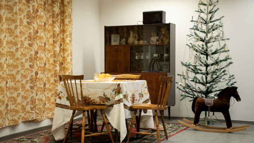 Fotografia z wystawy. W kadrze widać starą meblościankę na wysoki połysk, krzesła, stół z obrusem, tkaninę w kwietny wzór, konia na biegunach i sztuczną choinkę