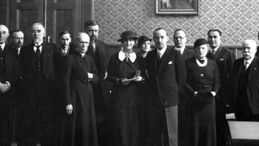 Obrazek do wydarzenia, czarno-biała fotografia grupy osób stojących w gabinecie pod obrazem marszałka Piłsudskiego. W centralnej części Kazimiera Iłłakowiczówna w kapeluszu.