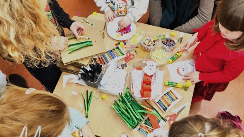 Obrazek do wydarzenia, widziany z góry stół, przy którym dzieci wykonują prace plastyczne z kolorowych materiałów.