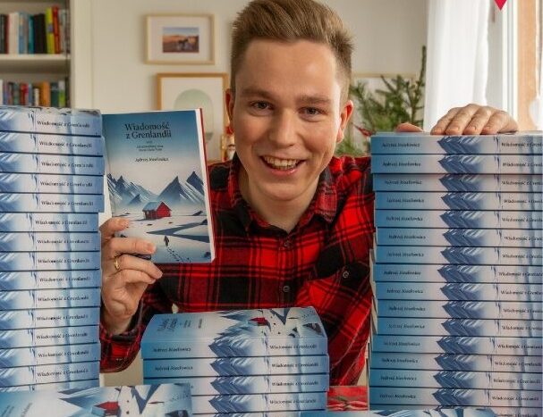 Obrazek do wydarzenia, autor, młody jasnowłosy mężczyzna w czerwonej kraciastej koszuli, obok równe stosy książek jego autorstwa