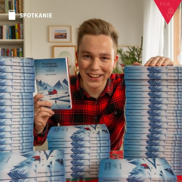 Obrazek do wydarzenia, autor, młody jasnowłosy mężczyzna w czerwonej kraciastej koszuli, obok równe stosy książek jego autorstwa
