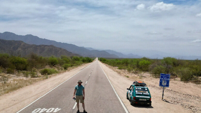 Obrazek do wydarzenia, asfaltowa droga na pustkowiu, na drodze namalowane liczby 4040, na środku mężczyzna w letnim ubraniu, obok turkusowy fiat maluch.