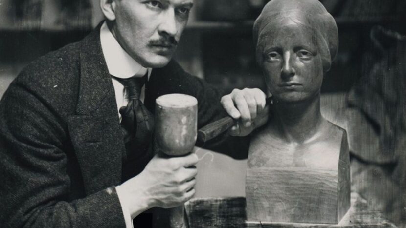 Obrazek do wydarzenia. Czarno biała fotografia artysty - mężczyzna w garniturze, z wąsem, patrzący w dal, trzyma dłuto przy rzeźbie - popiersiu kobiety.