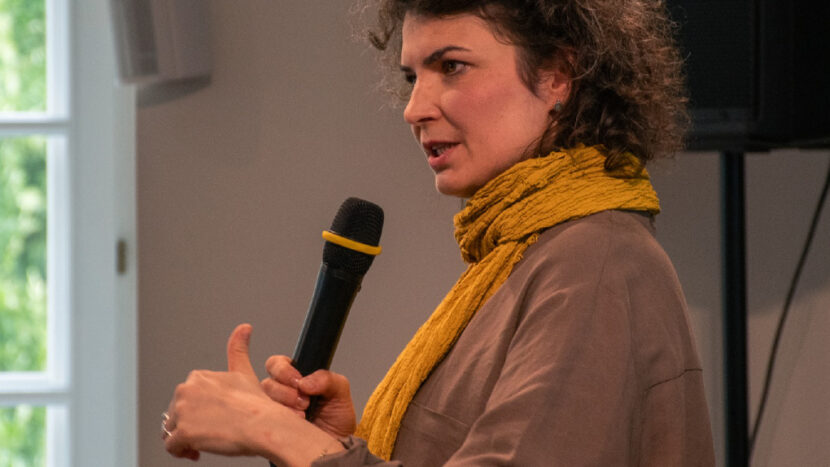 Obrazek do wydarzenia, kobieta w brązowym ubraniu i żółtym szalu, z kręconymi włosami, widoczna z profilu, trzyma mikrofon.
