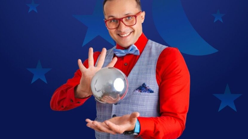 Obrazek do wydarzenia, na granatowym tle w gwiazdy mężczyzna w czerwonej koszuli i w okularach w czerwonej oprawce, ma uniesione ręce, między nimi zawieszona w powietrzu kula.