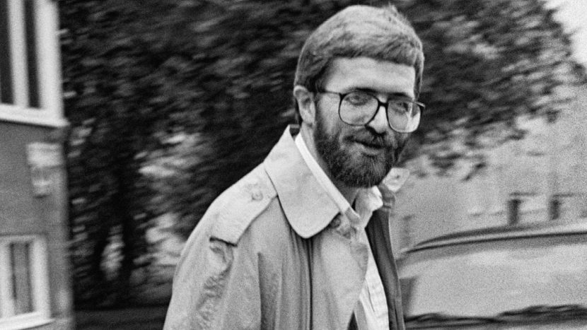 Obrazek do wydarzenia, czarno-biała fotografia Stanisława Barańczaka. Poeta, uśmiechnięty, w okularach i z zarostem idzie ulicą w rozpiętym płaszczu.